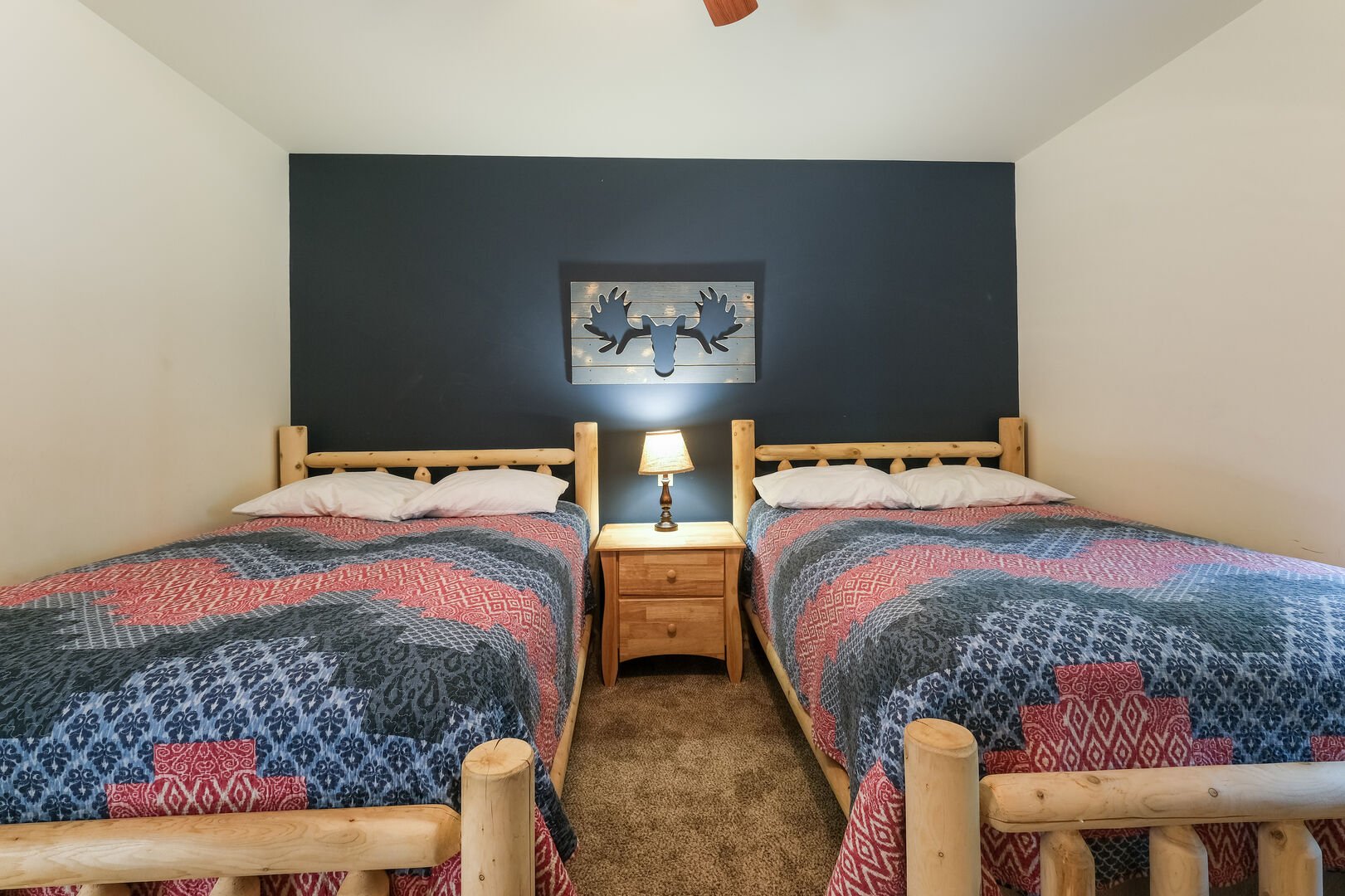 Two beds with nightstand in-between, a lamp on the nightstand shining onto a hanging moose wall decoration.
