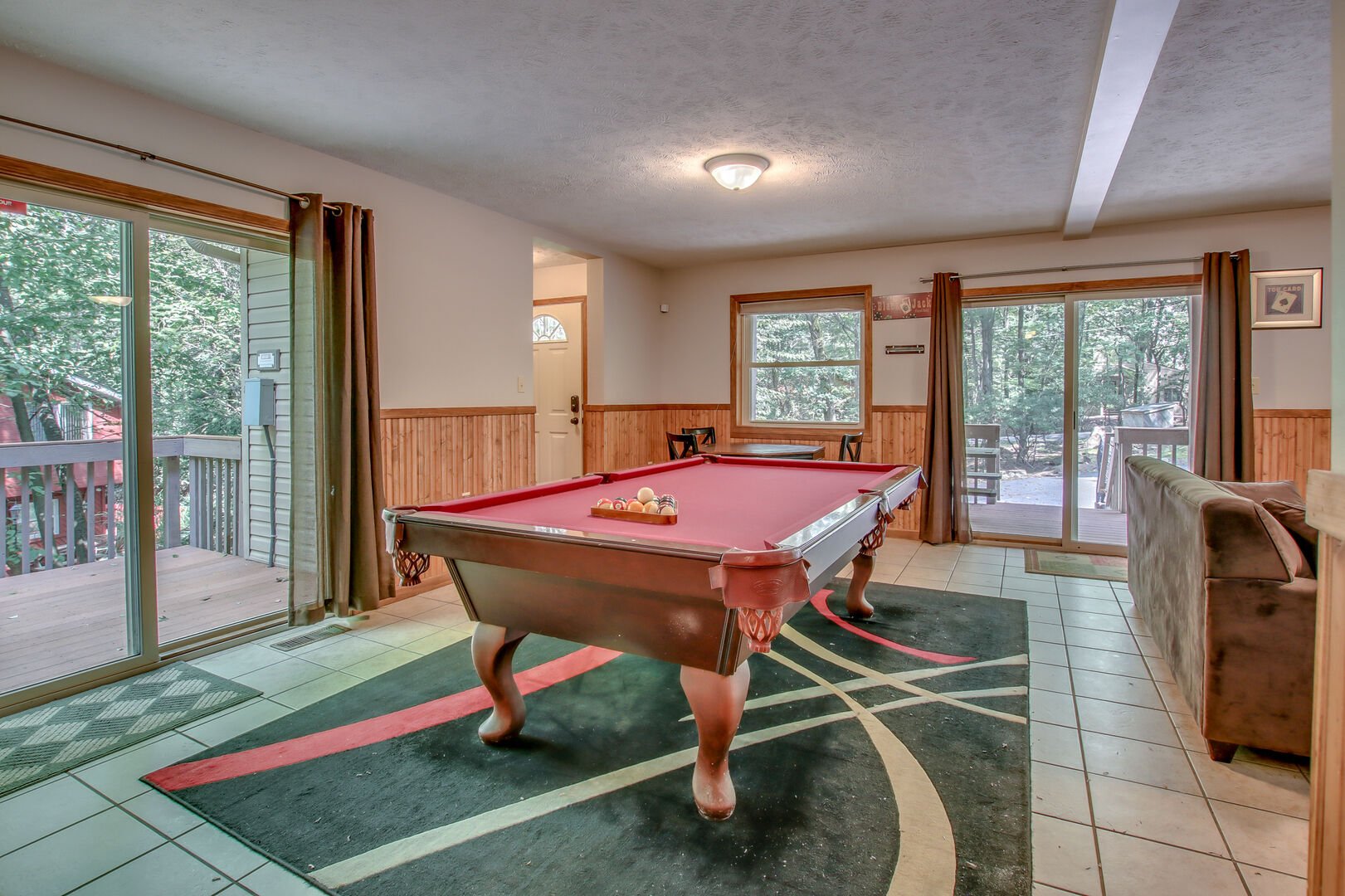 A pool table in the middle of the game room on a multicolored rug, with a couch to the right and sliding glass door to the left and behind.