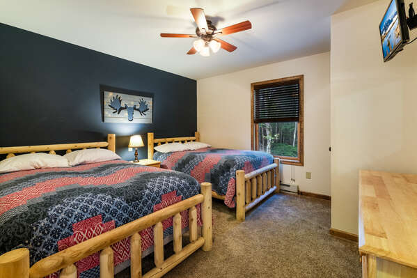 Two side-by-side beds, with a nightstand on either side, and a moose painting over them. Dresser and TV at foot of beds.