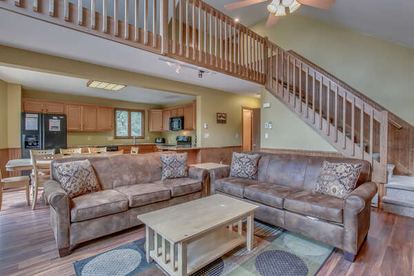 A picture of the great room, with two couches and a coffee table, with the kitchen and staircase in the background.