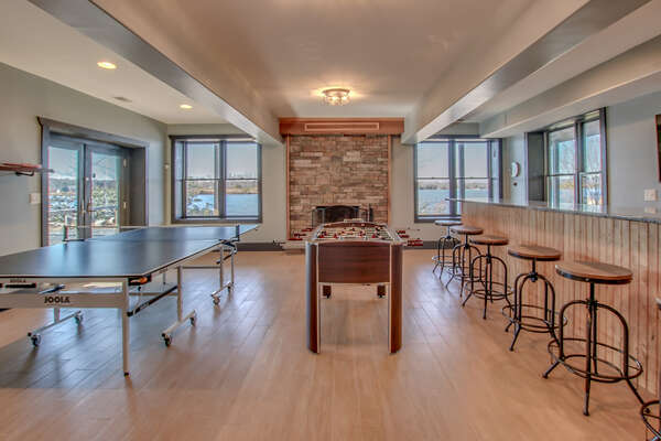 The game room with Foosball table, bar stools against the right wall, and ping pong table by the sliding glass door on the left. Fireplace in back.