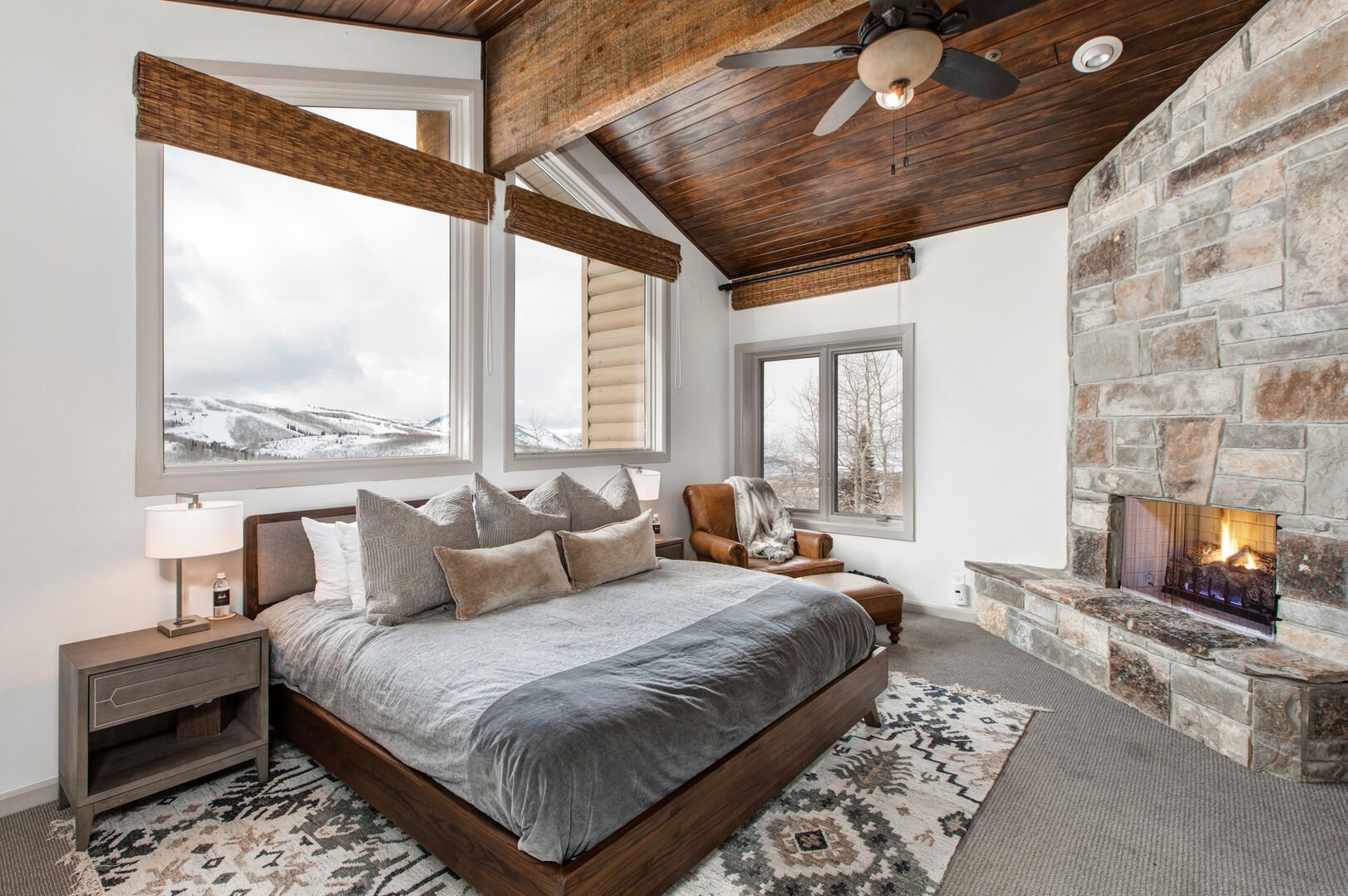 Upper level master bedroom. Custom-upholstered king bed, floor-to-ceiling stone fireplace, vaulted wooden plank ceiling, leather reading chair. Large picture windows show off gorgeous western views toward Deer Valley ski runs blanketed in snow.