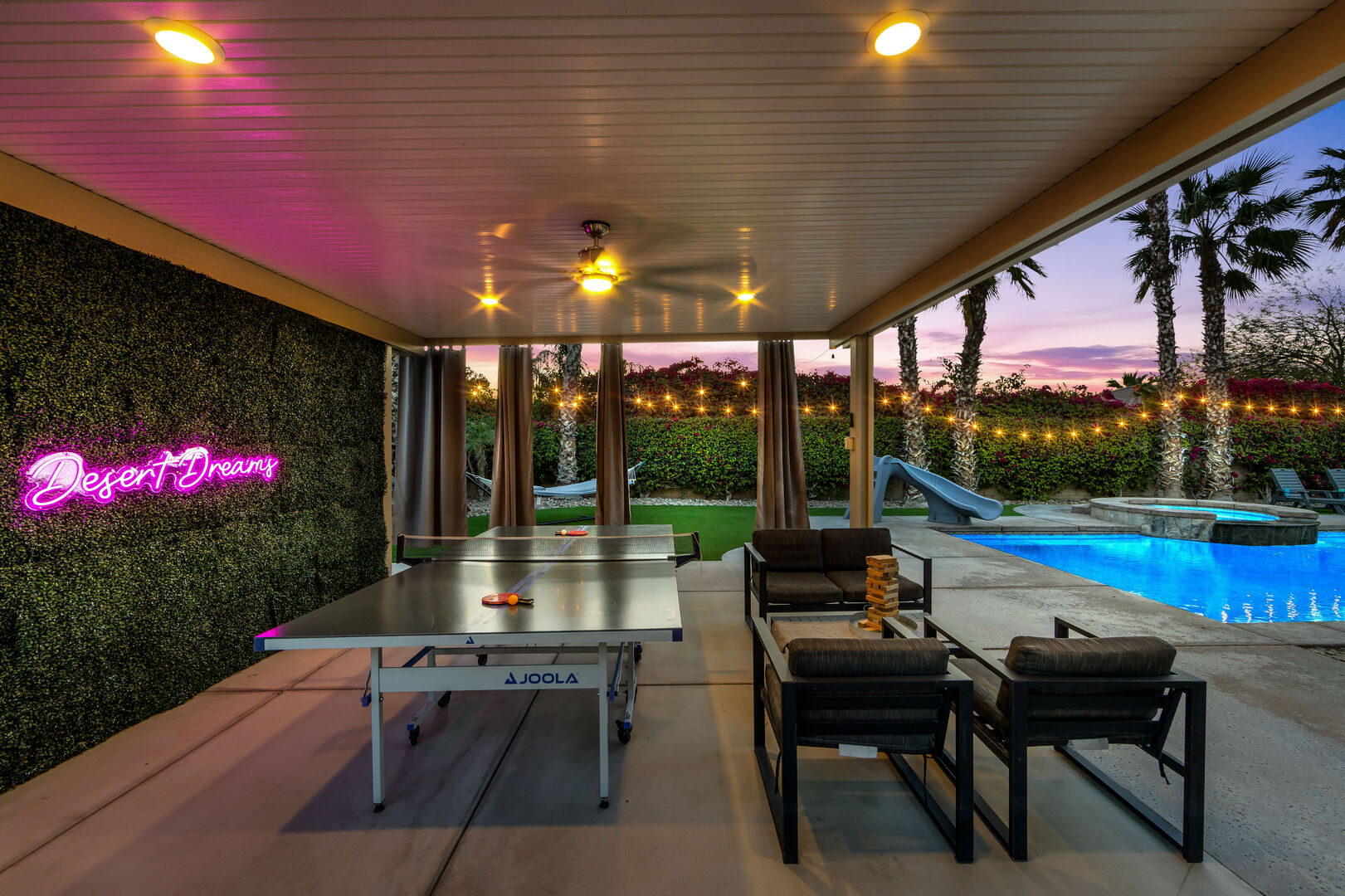 The shaded outdoor area features a ping pong table, an overhead fan with lighting, and a selfie-worthy faux background adorned with a light-up neon sign.
