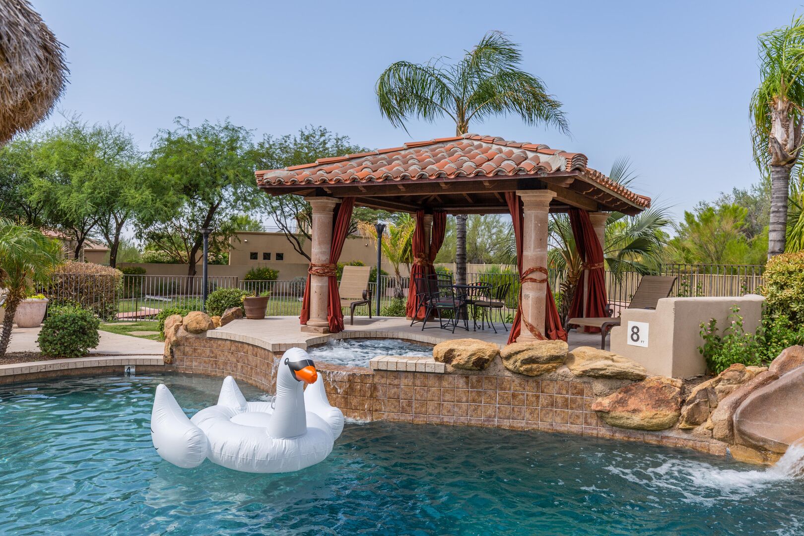 Tropical resort style backyard featuring a heated sparkling blue pool with a waterfall & built-in bar, a hot tub, covered terrace with outdoor dining, fire pit, and a lawn area for yard games surrounded by gorgeous mountain views.