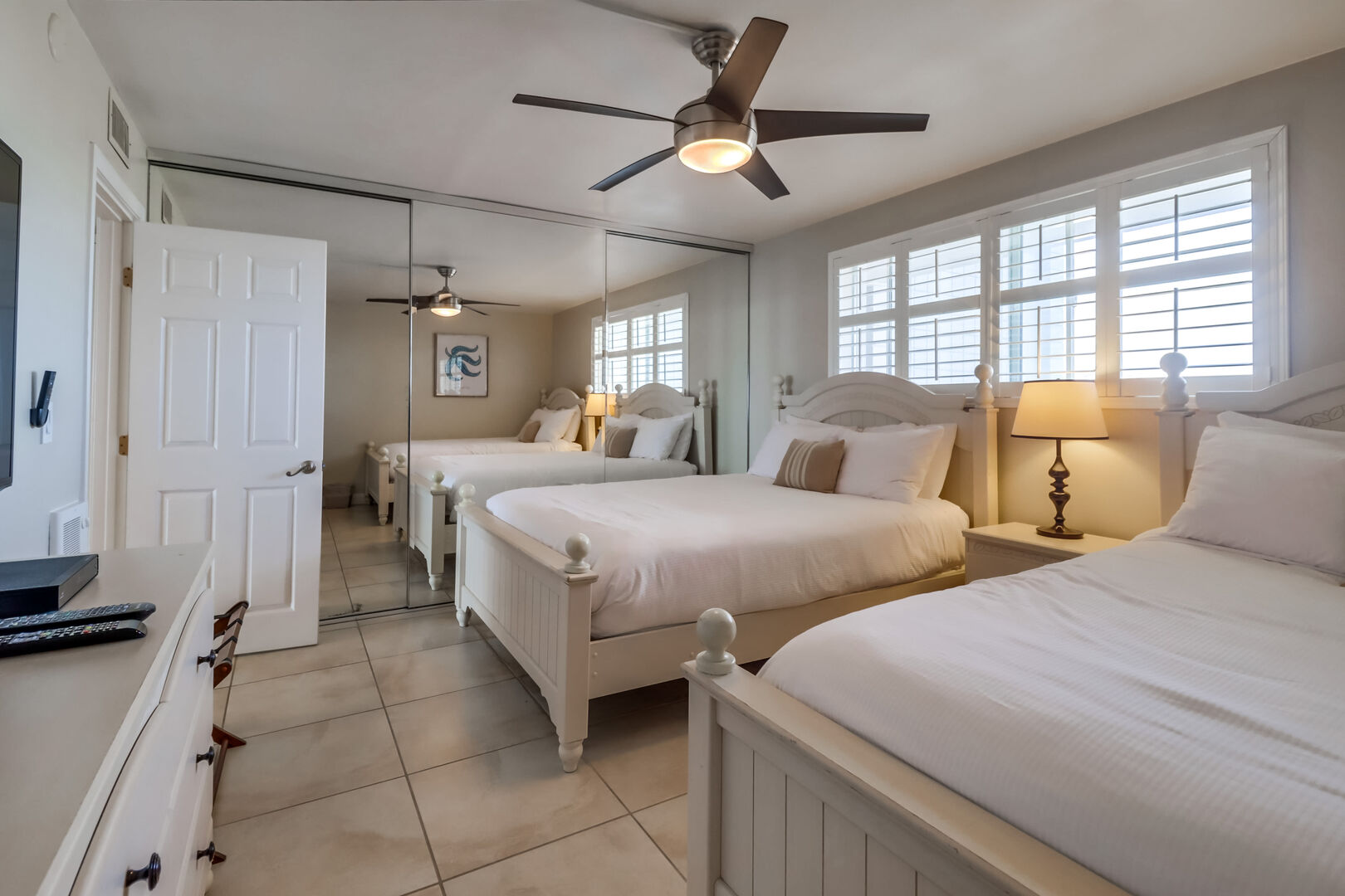 Clean and minimalist bedroom with 2 queen beds, ceiling fan, wall heater, dresser storage, luggage racks, large mirrored closet and smart TV with streaming capabilities