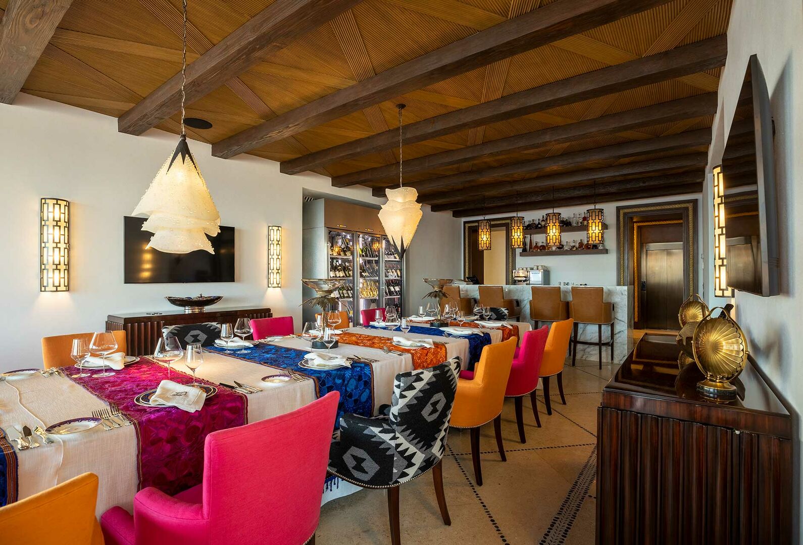 The long table of the dining area of La Datcha is filled with colorful chairs and a wall-mounted TV.