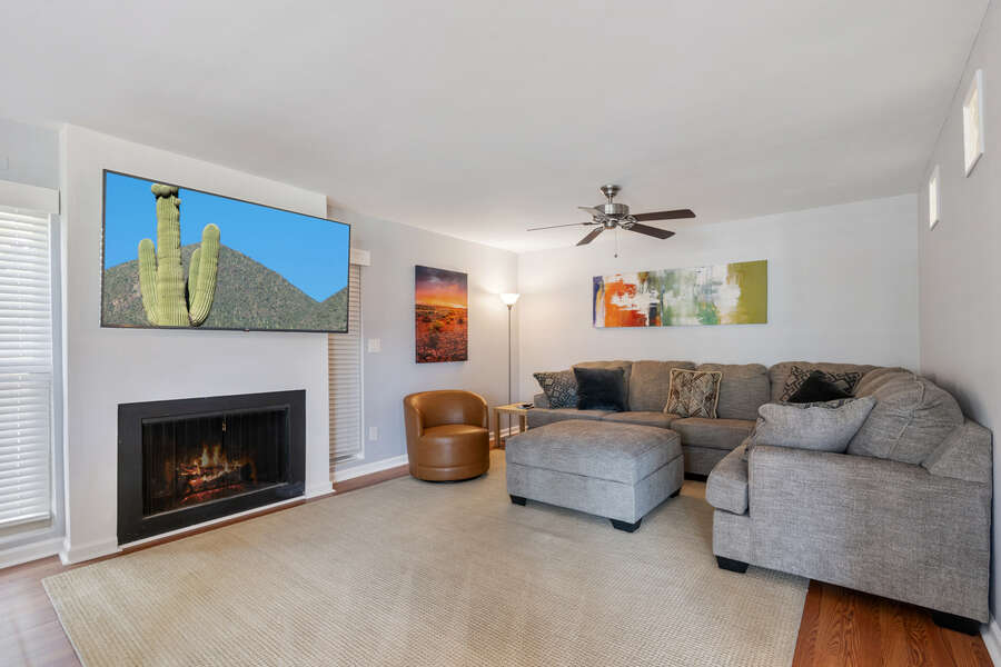 The cozy couch sits below a ceiling fan and in front of the living room fire place and large Smart TV.