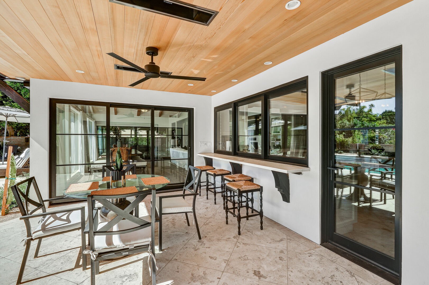 The outdoor dining area, equipped with ceiling heaters and bar stools, is located just outside the sliding kitchen window.