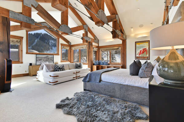 Large windows sit in front of the bed of the primary bedroom, with a large couch in front of an entertainment center and TV.