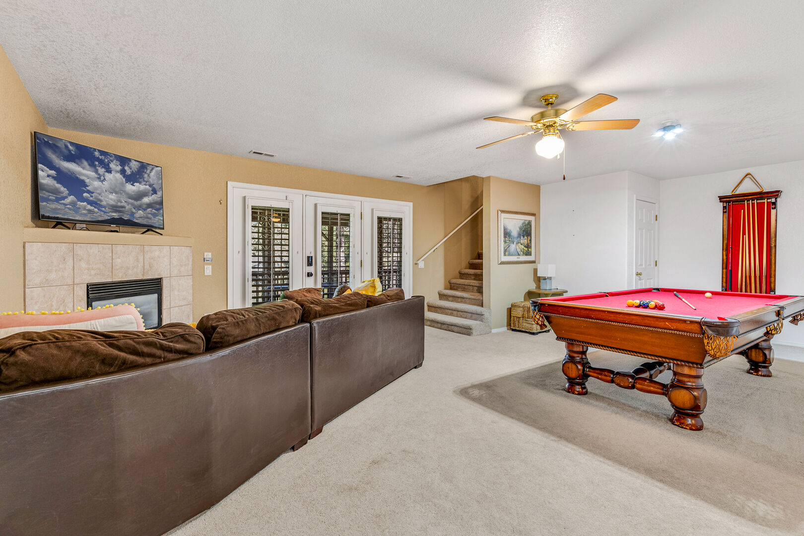 This inviting entertainment room downstairs features a cozy seating area with a brown leather sectional, a mounted TV above a fireplace, and a stylish red-felt pool table.